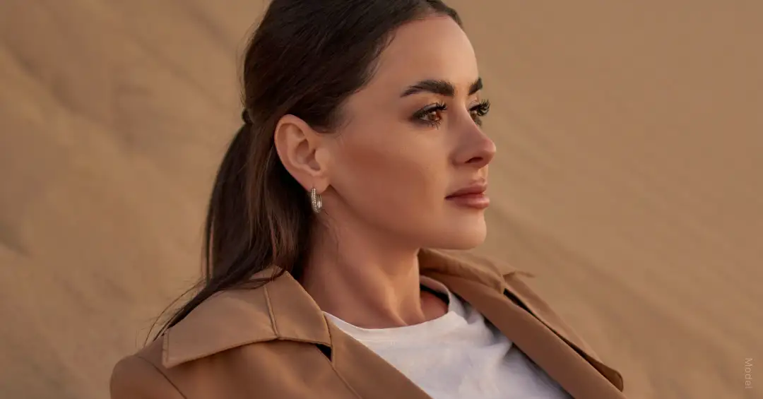 Woman sitting in sand wearing a brown jacket and white shirt gazing out